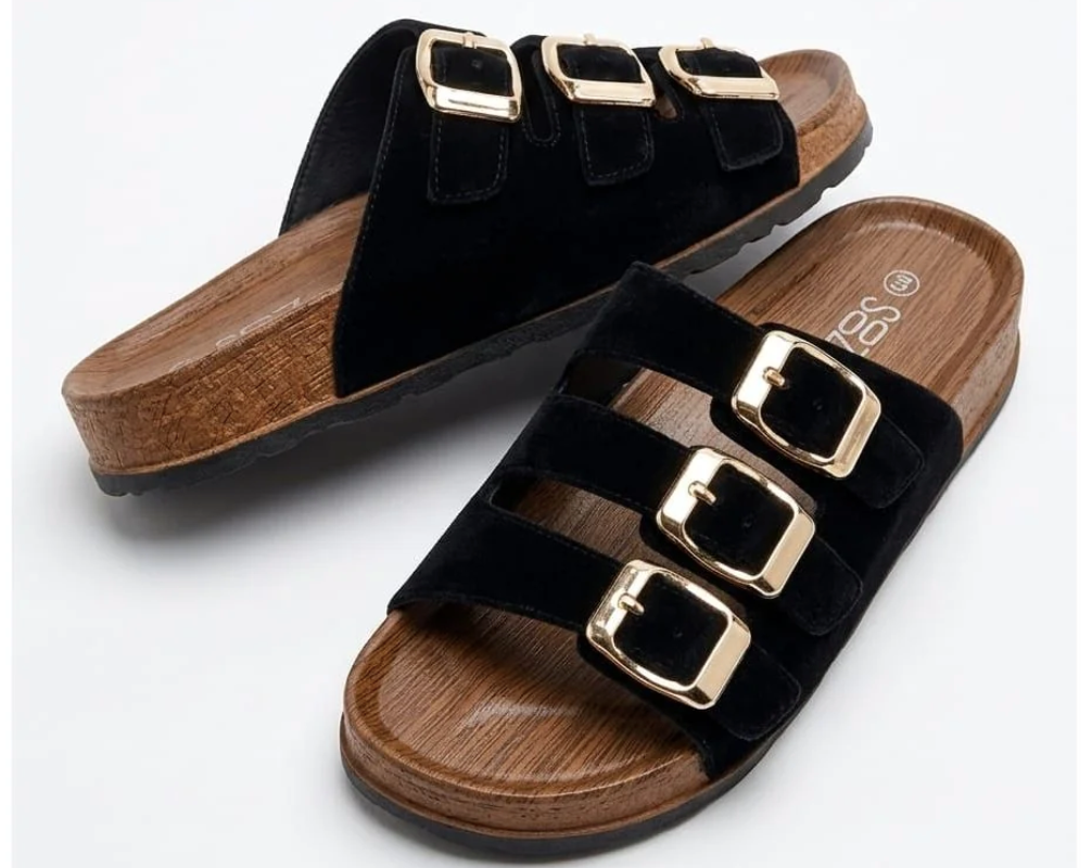 A pair of black velvet slide sandals with three gold-tone buckle straps and a brown cork-textured footbed, shown on a light gray background.